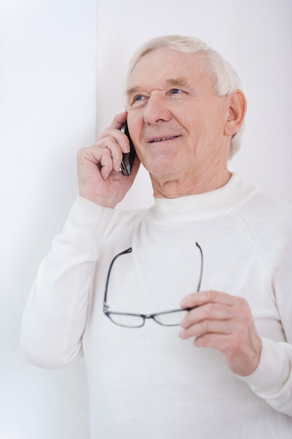 Usando aparelhos modernos. Homem sênior alegre falando ao celular e segurando os óculos enquanto se encosta na parede