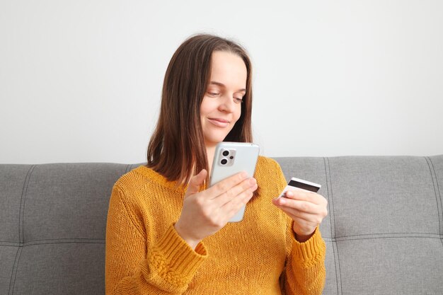 Foto usando carteira entrando transação de compra digital mulher caucasiana de cabelos castanhos vestindo suéter laranja usando telefone celular e segurando cartão de crédito enquanto está sentada no sofá em casa