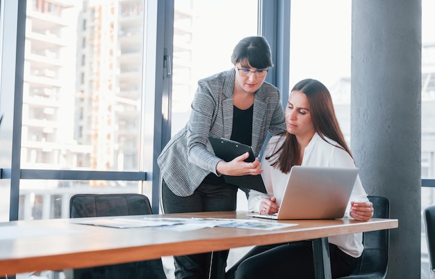 Usando una computadora portátil Dos mujeres vestidas formalmente están adentro en la oficina moderna trabajando juntas