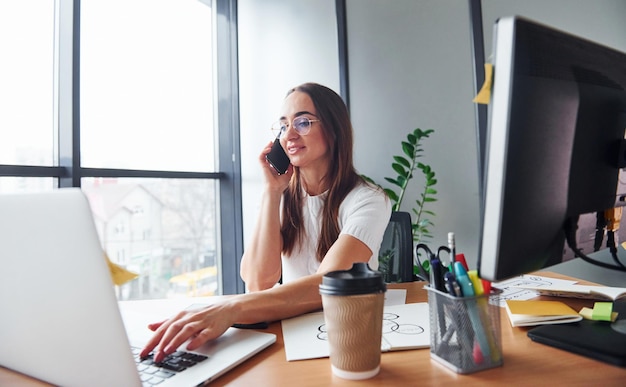 Usando una computadora portátil, una mujer adulta joven con ropa formal está adentro en la oficina