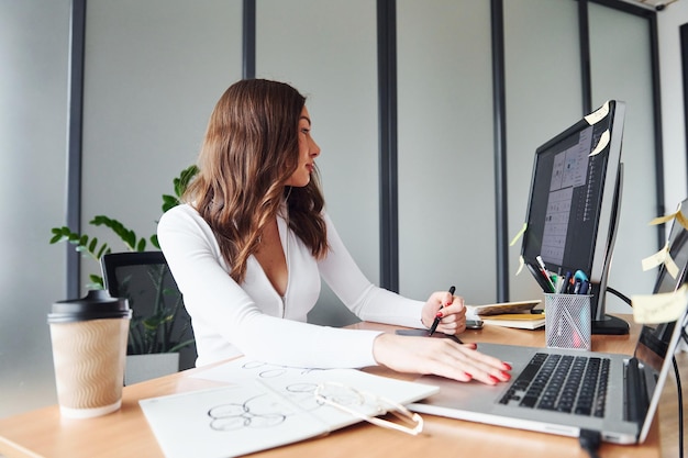 Usando una computadora portátil, una mujer adulta joven con ropa formal está adentro en la oficina