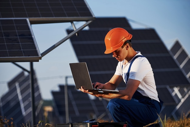 Usando computadora portátil. Trabajador de sexo masculino en uniforme azul al aire libre con baterías solares en un día soleado.