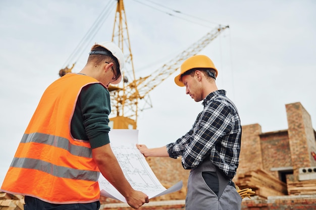 Usando documento em papel com plano Dois trabalhadores da construção civil em uniforme e equipamentos de segurança têm trabalho na construção juntos