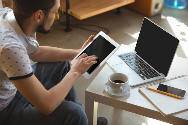 Usando gadgets. Hombre trabajando desde casa durante la cuarentena por coronavirus o COVID-19, concepto de oficina remota. Joven empresario, gerente haciendo tareas con teléfono inteligente, computadora, conferencia en línea, reunión.