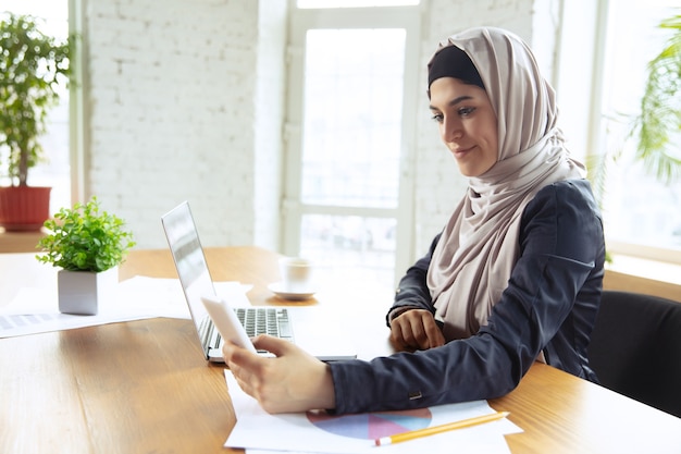 Usando gadgets. retrato de uma linda mulher de negócios árabe usando hijab enquanto trabalhava no openspace ou escritório. conceito de ocupação, liberdade na área de negócios, liderança, sucesso, solução moderna.