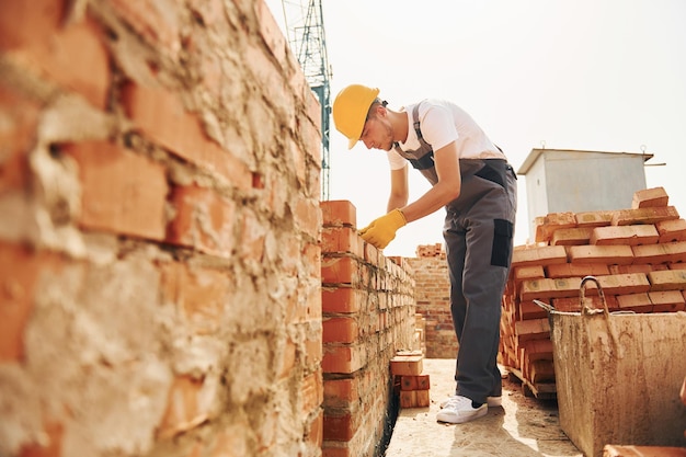 Usando ladrillos, el joven trabajador de la construcción en uniforme está ocupado en el edificio sin terminar