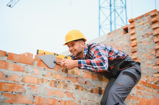 Usando ladrillos, el joven trabajador de la construcción en uniforme está ocupado en el edificio sin terminar