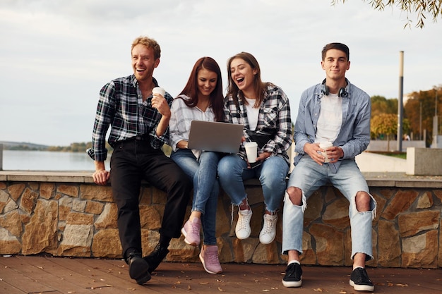 Usando laptop Grupo de jovens amigos alegres que estão ao ar livre se divertindo juntos