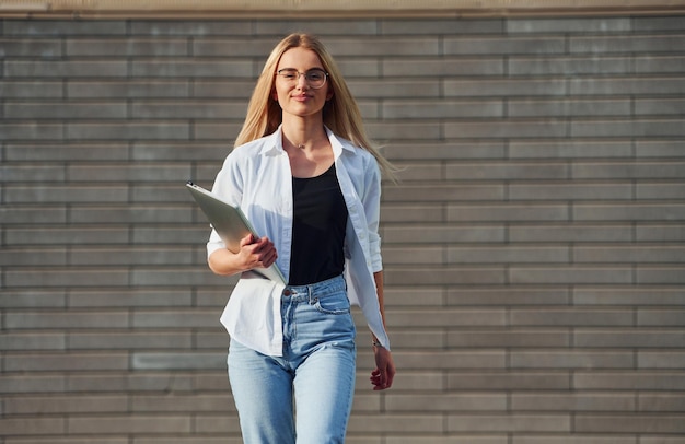 Usando una laptop Hermosa rubia con ropa informal está al aire libre durante el día soleado