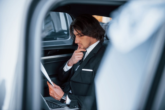 Usando o bloco de notas. Jovem empresário de terno preto e gravata dentro de um automóvel moderno.