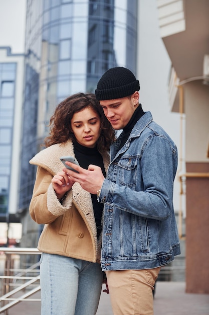 Usando o telefone Casal alegre em roupas quentes casuais caminha ao ar livre na cidade perto do prédio comercial