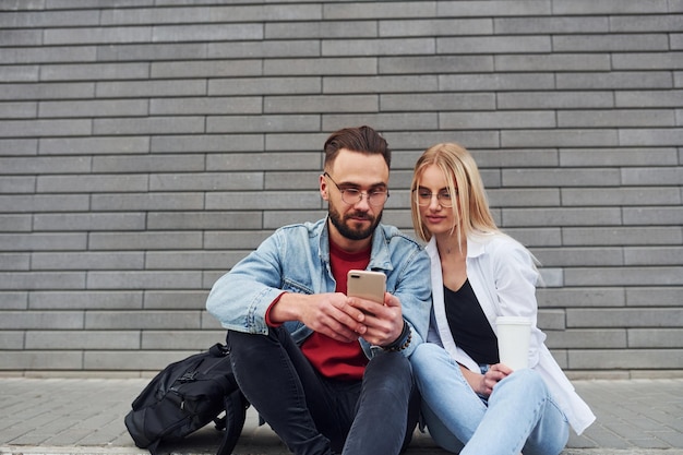 Usando o telefone Jovem elegante com mulher em roupas casuais sentados ao ar livre juntos Concepção de amizade ou relacionamentos