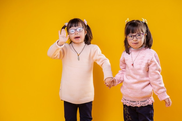 Usando óculos transparentes. garotas fofas sorridentes com anormalidade cromossômica ficando juntas e acenando com as mãos em um gesto de boas-vindas