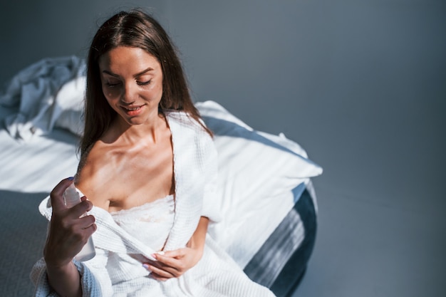 Usando perfume. Retrato da bela morena com roupas brancas que se senta na cama na sala. Boa iluminação.