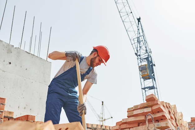 Usando un showel Joven trabajador de la construcción en uniforme está ocupado en el edificio sin terminar