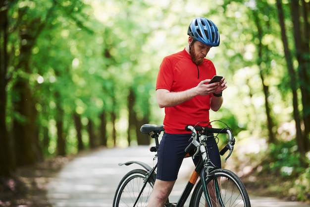 Usando smartphone. Ciclista de bicicleta está na estrada de asfalto na floresta em um dia ensolarado