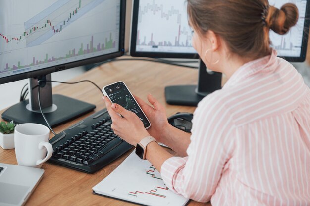 Foto usando smartphone corretor de ações feminino em roupas casuais está trabalhando no escritório pelo pc