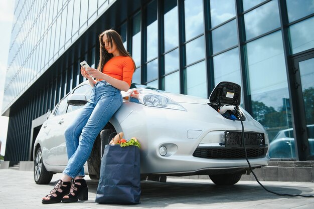 Usando smartphone enquanto espera Mulher na estação de carga de carros elétricos durante o dia Veículo novo em folha