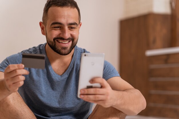 Foto usando smartphone guapo barbudo con tarjeta de débito o crédito en manos comprando en línea.