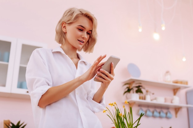 Usando smartphone. Mulher atraente com cabelo curto em pé na cozinha e usando smartphone