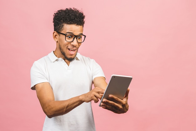 Usando tablet. Homem africano bonito jovem sorrindo segurando o tablet e jogando jogos ou usando um aplicativo de reserva isolado contra parede rosa.