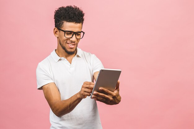 Usando tableta. Hombre africano hermoso joven que sonríe sosteniendo la tableta y jugando juegos o usando una aplicación de reserva aislada contra la pared rosada.