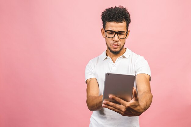 Usando tableta. Hombre africano hermoso joven que sostiene la tableta y que juega a juegos o que usa una aplicación de reserva aislada contra la pared rosada.