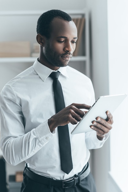 Usando tecnologias modernas. Jovem africano bonito em trajes formais trabalhando em um tablet digital enquanto está no escritório de criação