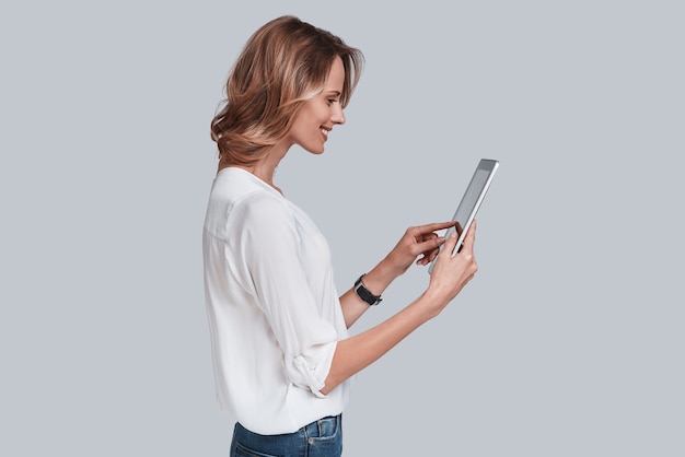 Usando tecnologias modernas. Mulher jovem e atraente segurando um tablet digital e olhando para ele em pé contra um fundo cinza