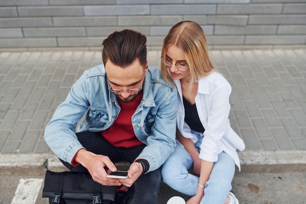 Usando el teléfono Hombre elegante joven con mujer en ropa casual sentados juntos al aire libre Concepción de amistad o relaciones