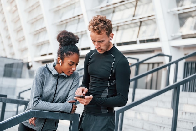 Usando el teléfono, un hombre europeo y una mujer afroamericana con ropa deportiva hacen ejercicio juntos