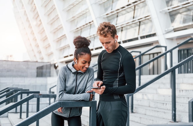 Usando el teléfono, un hombre europeo y una mujer afroamericana con ropa deportiva hacen ejercicio juntos