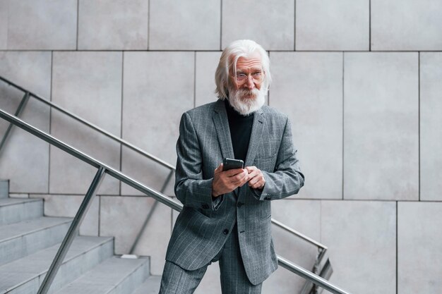 Usando un teléfono inteligente Un hombre de negocios mayor vestido con ropa formal con cabello gris y barba está al aire libre