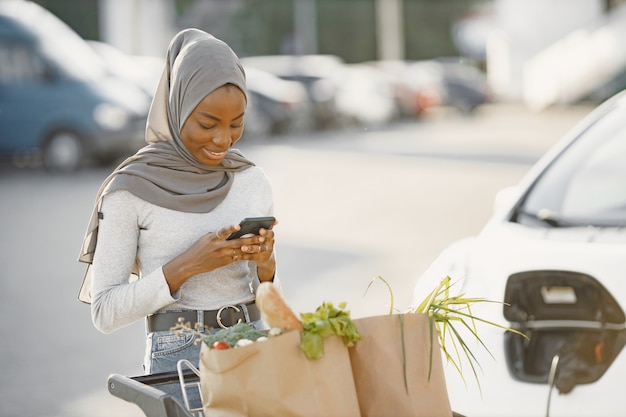 Usando un teléfono inteligente mientras espera. Mujer de etnia africana en la estación de carga de coches eléctricos durante el día. Vehículo nuevo a estrenar.