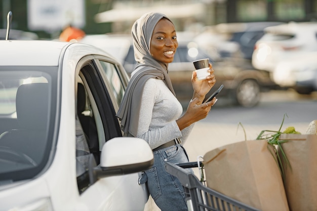 Usando un teléfono inteligente mientras espera. Mujer de etnia africana en la estación de carga de coches eléctricos durante el día.