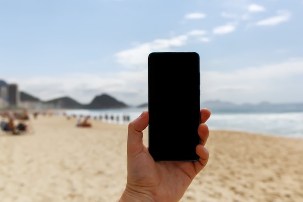 Usando un teléfono inteligente en la playa. Pantalla negra en blanco para su contenido.