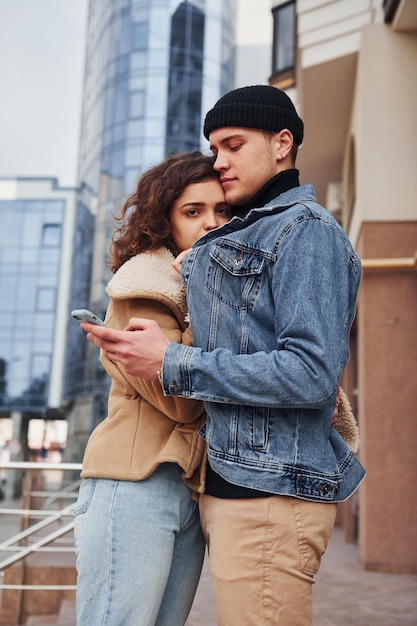 Usando el teléfono Una pareja alegre con ropa informal de abrigo da un paseo al aire libre en la ciudad cerca del edificio de negocios