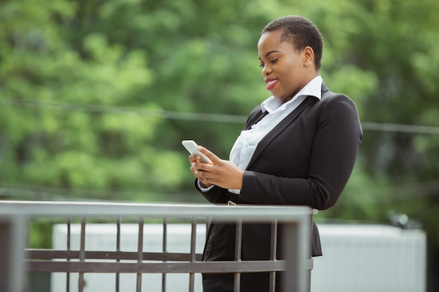 Usando um smartphone, uma mulher de negócios afro-americana em traje de escritório sorrindo parece confiante