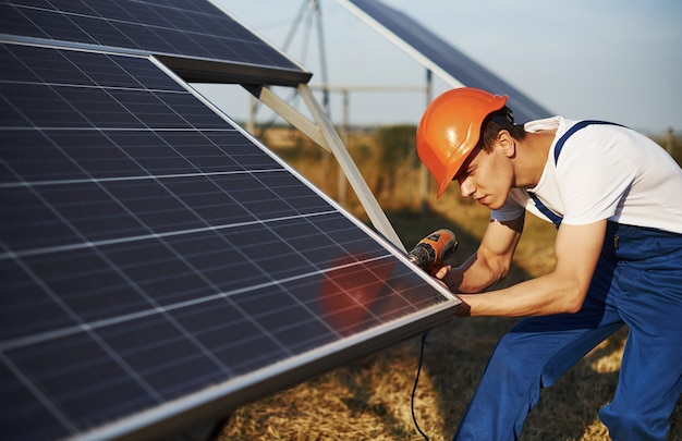 Usando uma chave de fenda sem fio. Trabalhador masculino com uniforme azul ao ar livre com baterias solares em dia ensolarado.