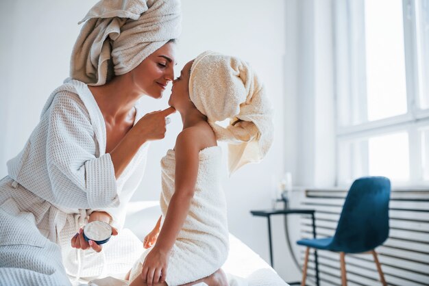 Usar creme para limpar a pele. Jovem mãe com sua filha tem dia de beleza dentro de casa, no quarto branco.