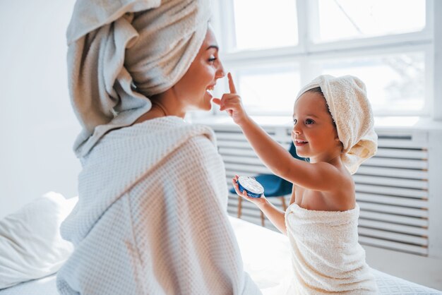 Usar creme para limpar a pele. Jovem mãe com sua filha tem dia de beleza dentro de casa, no quarto branco.