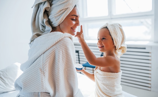 Usar creme para limpar a pele. Jovem mãe com sua filha tem dia de beleza dentro de casa, no quarto branco.
