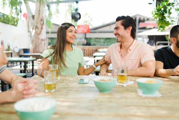 Usted es tan divertido. Hermosa joven y hombre hispano coqueteando con bebidas mientras hablan durante una reunión con un grupo de amigos en un bar hipster