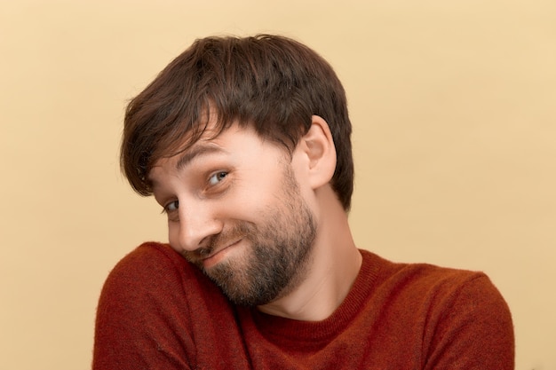 Usted me hace sonrojar. Foto de un joven tímido con barba con un suéter, se da la vuelta y mira por debajo de las cejas, posando contra la pared beige