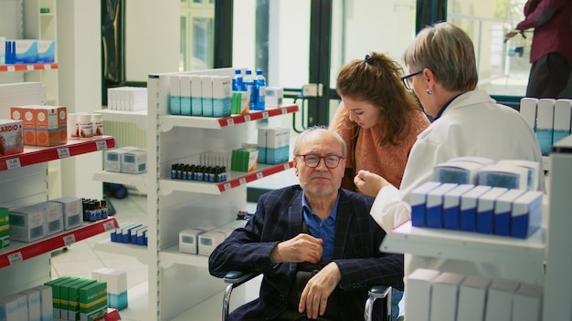 La usuaria de silla de ruedas y la cuidadora entregan papel de prescripción al especialista y piden comprar pastillas para el tratamiento. Joven cuidador que ayuda a un adulto con discapacidad crónica en una farmacia. Disparo de trípode.