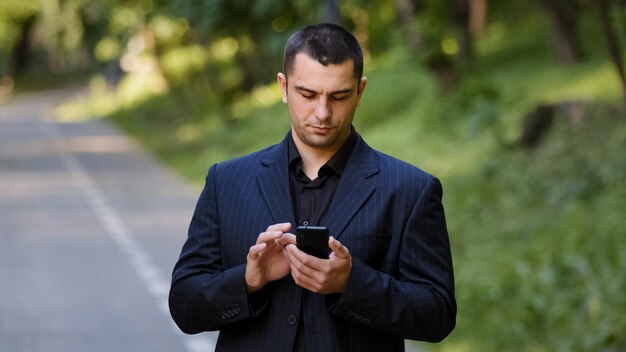 Foto el usuario adulto serio del hombre caucásico está parado al aire libre en el parque viste traje formal negro y sostiene el móvil