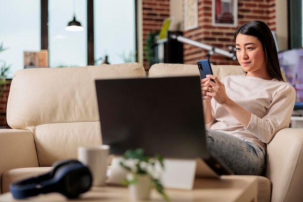 Usuário de smartphone de mulher asiática em casa no sofá, olhando para o telefone com um sorriso autêntico. usuário móvel da tecnologia da web da internet