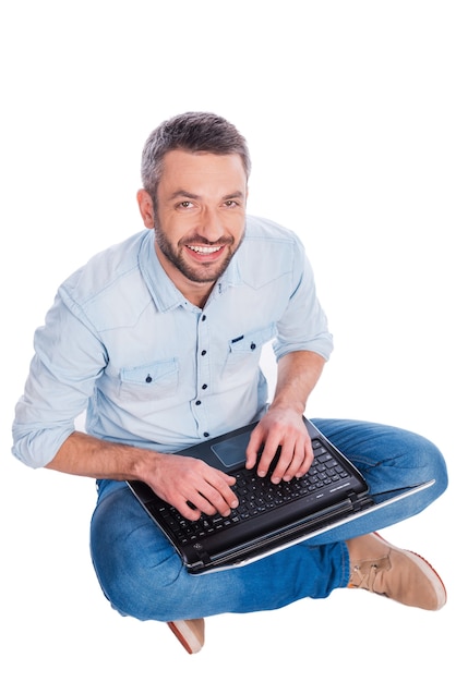 Usuario feliz. Vista superior del apuesto joven en ropa casual sentado en el suelo y trabajando en la computadora portátil mientras está aislado sobre fondo blanco.