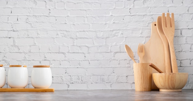 Utensilios de cocina y tazas de madera sobre fondo de textura de pared de ladrillo blanco.