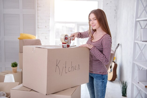 Utensilios necesarios. Bastante joven posando para la cámara mientras empaca utensilios de cocina y cierra la caja con cinta adhesiva